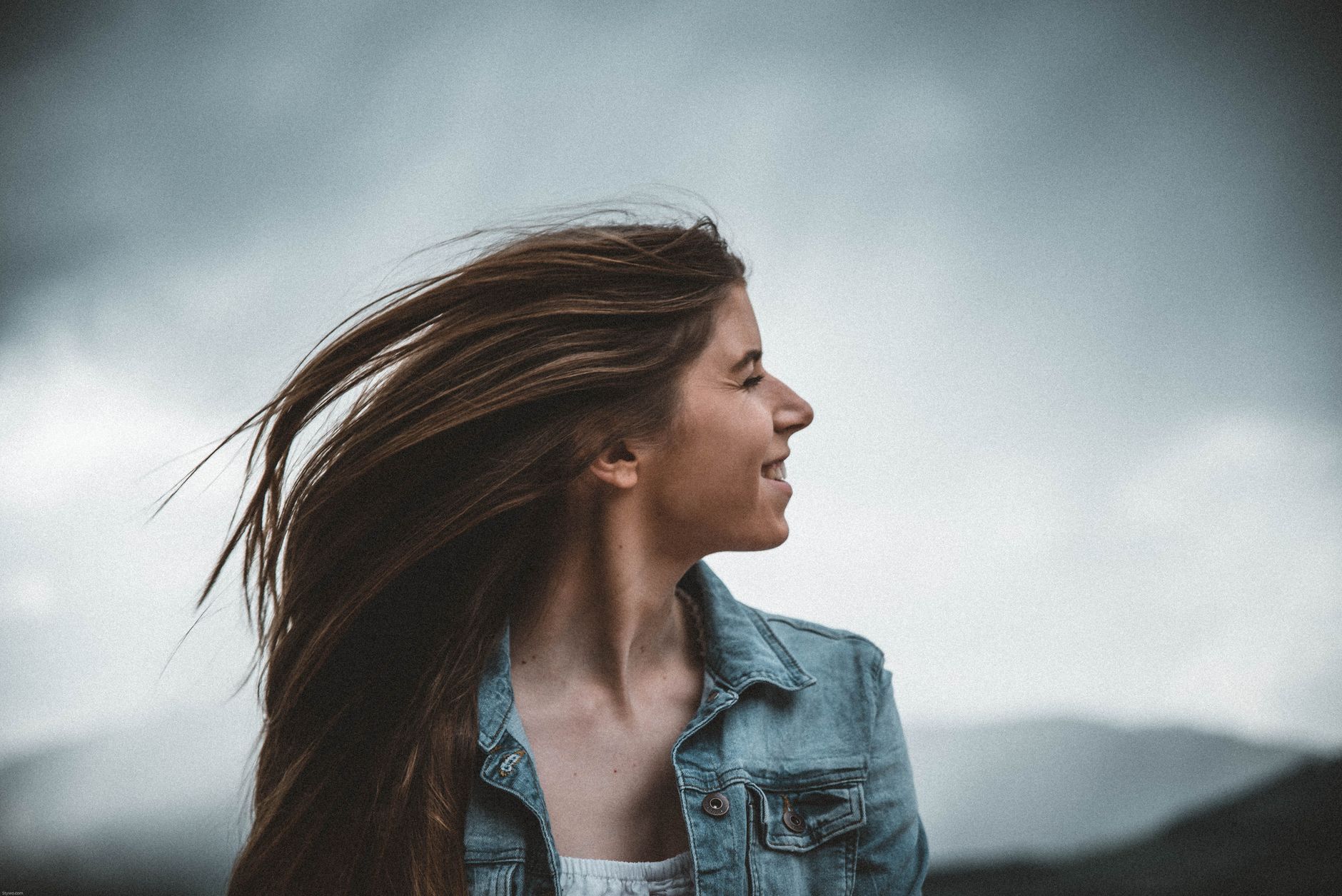 Porträt einer lächelnden jungen Frau in einer Jeansjacke vor einem bewölkten Himmel.