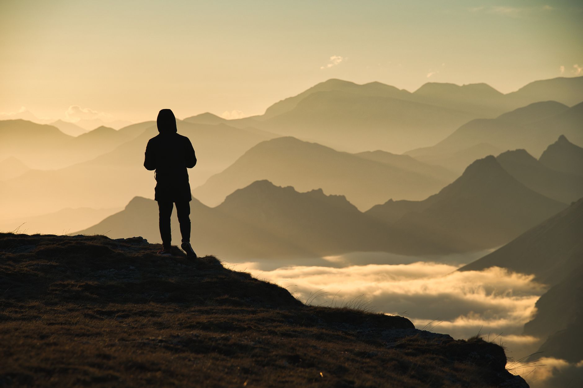 Silhouette einer Person, die auf einer Klippe steht mit atemberaubendem Bergblick bei Sonnenaufgang, umgeben von Wolken.