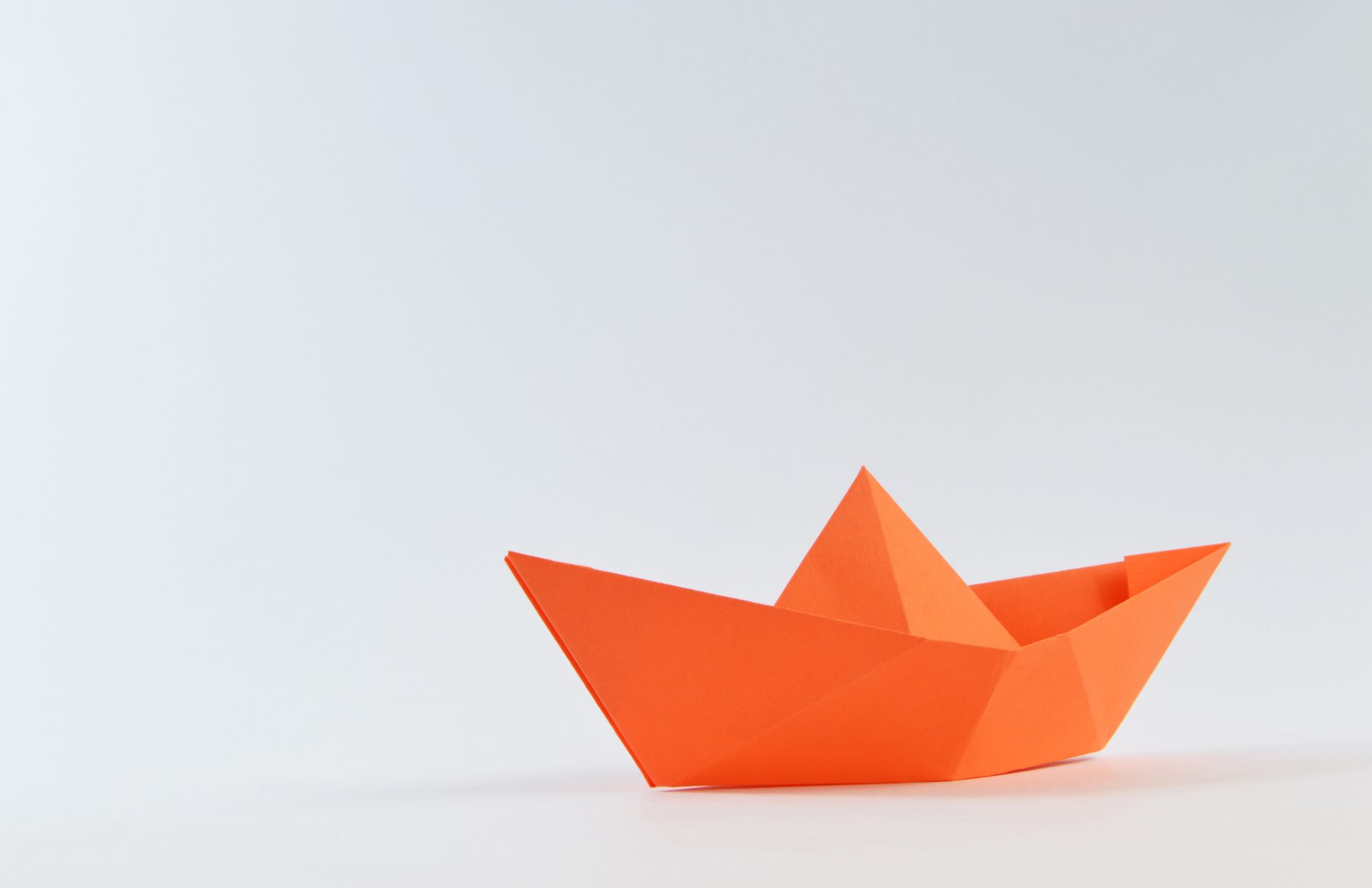 Minimalistisches Bild eines orangen Origami-Papierboots auf einem weißen Hintergrund, perfekt für Kreativität und Bastelkonzepte.